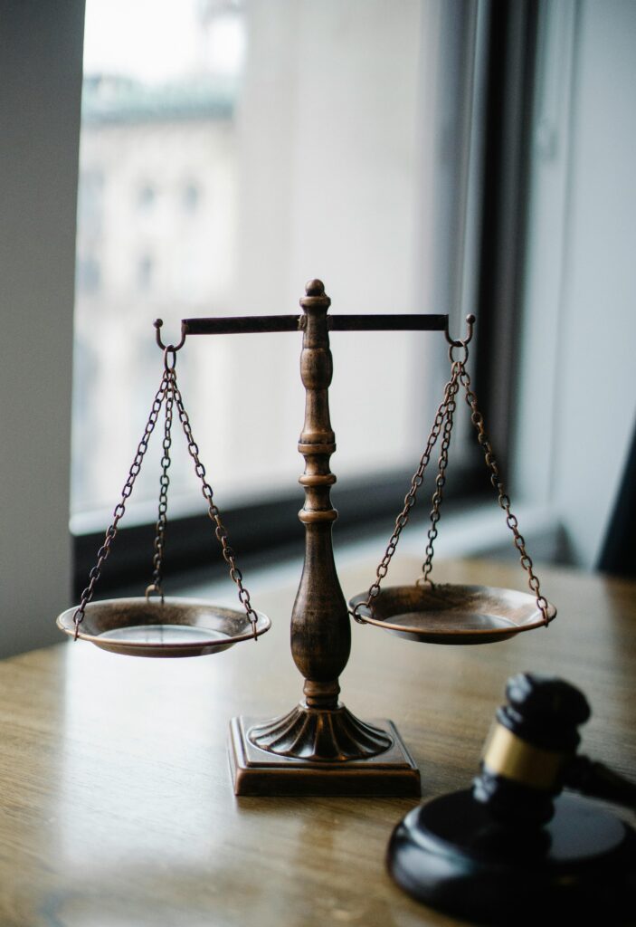 Balanced justice scale on a desk next to a judge’s gavel with a blurred window background